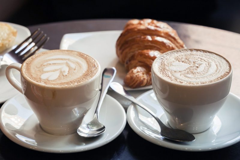 Dos tazas de café con espuma de leche y un cruasán sobre una mesa en una cafetería
